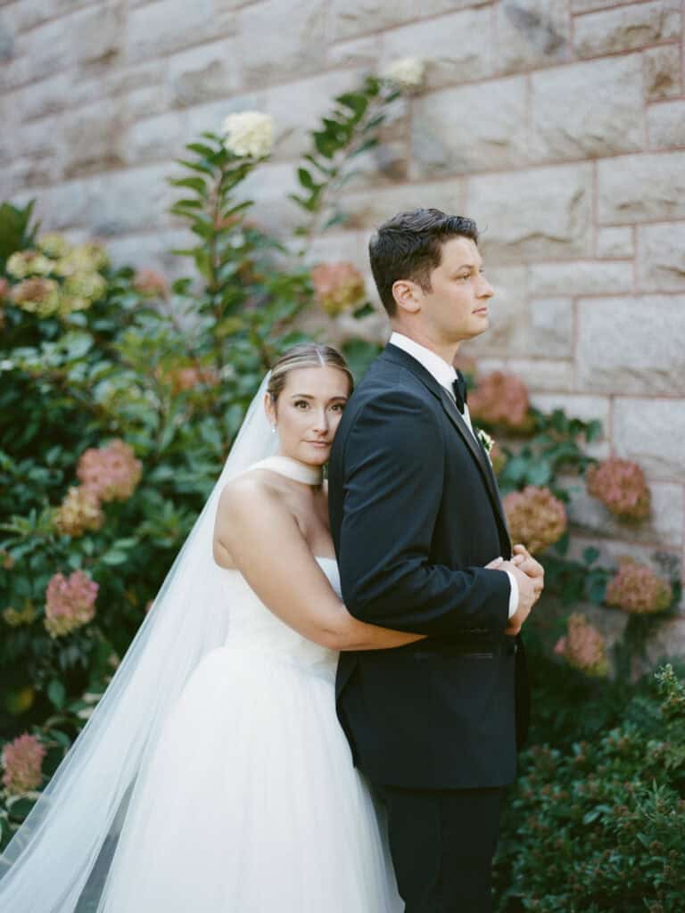 A bride in a white dress and veil embraces a groom in a black suit before a stone wall and greenery, captured in timeless elegance by Nantucket Wedding Photographer Kaity Farrell on film.