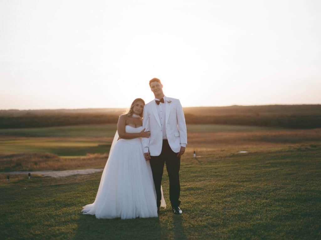 A bride in a white dress and a groom in a white tuxedo jacket stand together on a grassy field at sunset, captured in a timeless film photo by Nantucket Wedding Photographer Kaity Farrell.