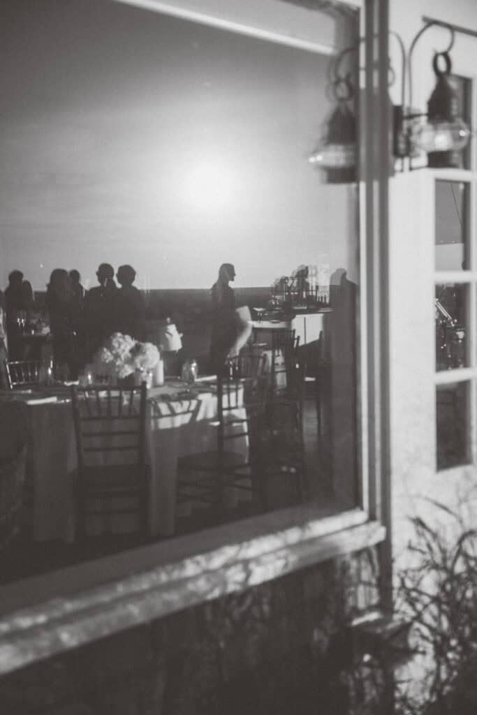 Black and white photo by Nantucket Wedding Photographer Kaity Farrell shows people and tables through a window, with reflections blending indoor and outdoor scenes at an event.