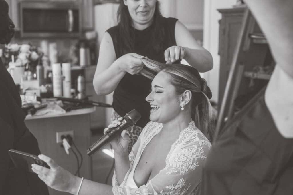 A woman in a lace robe smiles and sings into a microphone while getting her hair styled by another woman in a kitchen setting.
