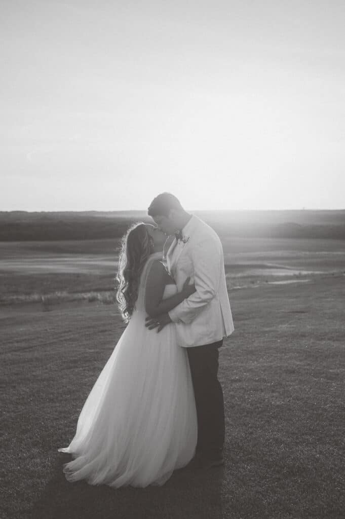 A bride and groom stand close together on a grassy field at sunset, facing each other, with the bride in a flowing dress and the groom in a light suit—captured beautifully by Nantucket Wedding Photographer Kaity Farrell.