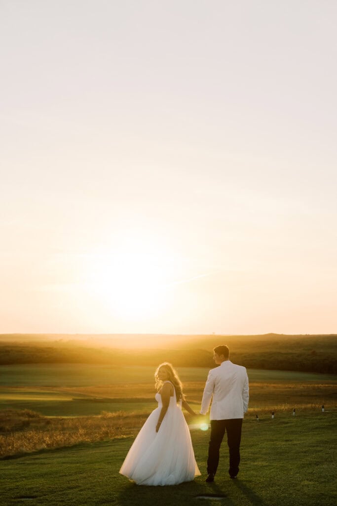 A bride and groom walk hand in hand on a grassy field at sunset, the sun low on the horizon and an open landscape behind them, perfectly captured by Nantucket Wedding Photographer Kaity Farrell.