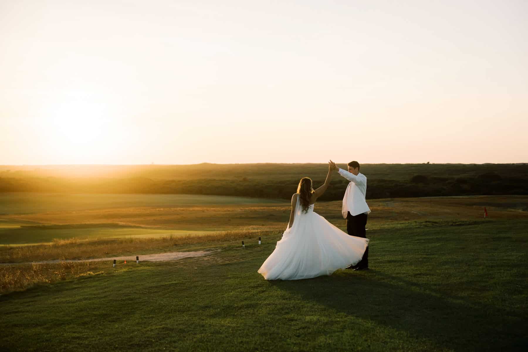 A couple dances on a grassy field at sunset, the woman in a white dress and the man in a white shirt and dark pants, beautifully captured by Nantucket wedding photographer Kaity Farrell as the sun dips low on the horizon.