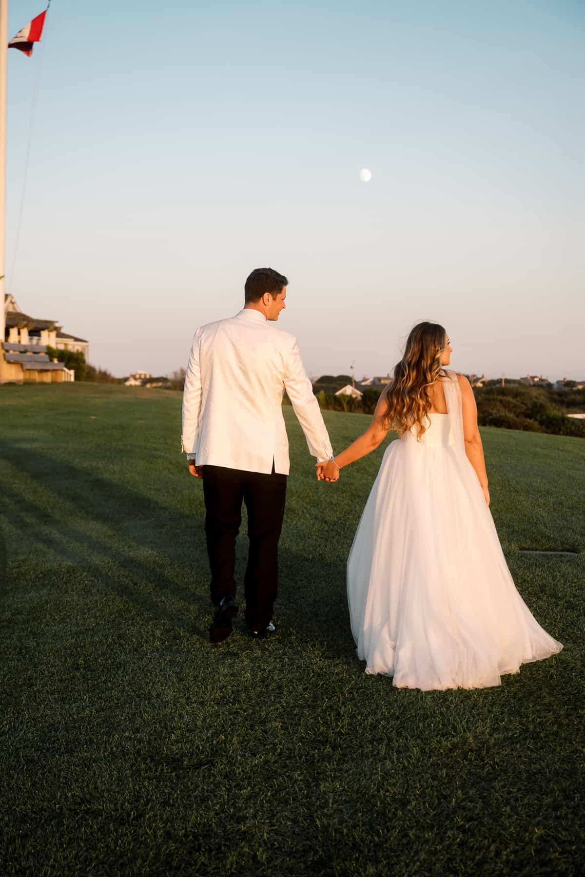 A bride and groom holding hands walk across a grassy lawn at sunset, with a clear sky and buildings visible in the background, beautifully captured by Nantucket Wedding Photographer Kaity Farrell.