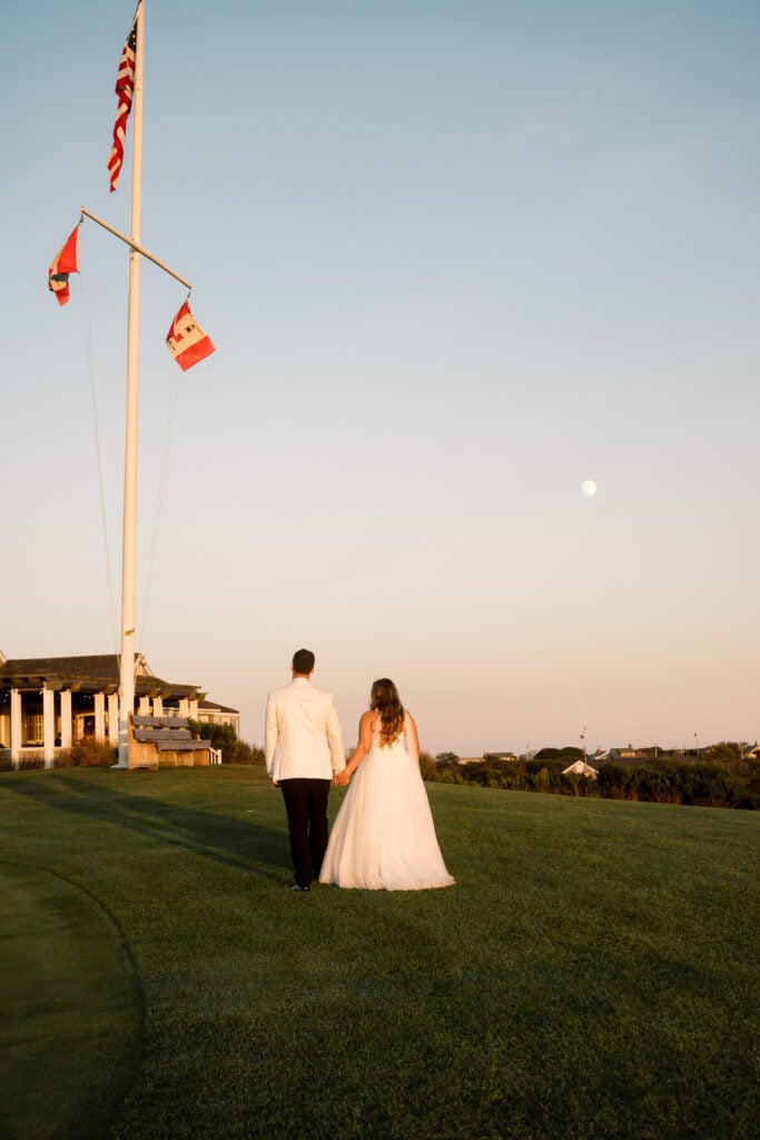 A bride and groom walk hand in hand on a grassy lawn at sunset, with flagpoles and a building in the background, beautifully captured by Nantucket Wedding Photographer Kaity Farrell.
