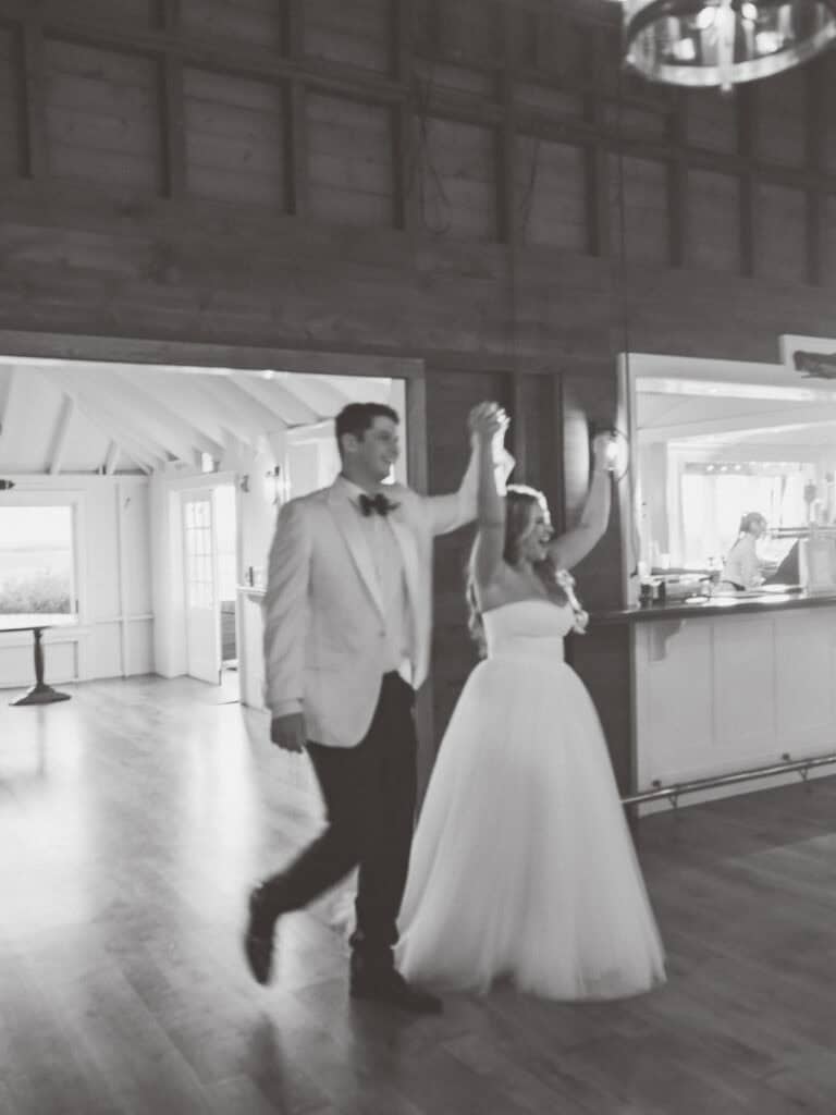 A bride and groom enter a reception hall, holding hands and raising their arms. Captured in black and white with a soft blur, this moving moment is artfully documented by Nantucket Wedding Photographer Kaity Farrell.