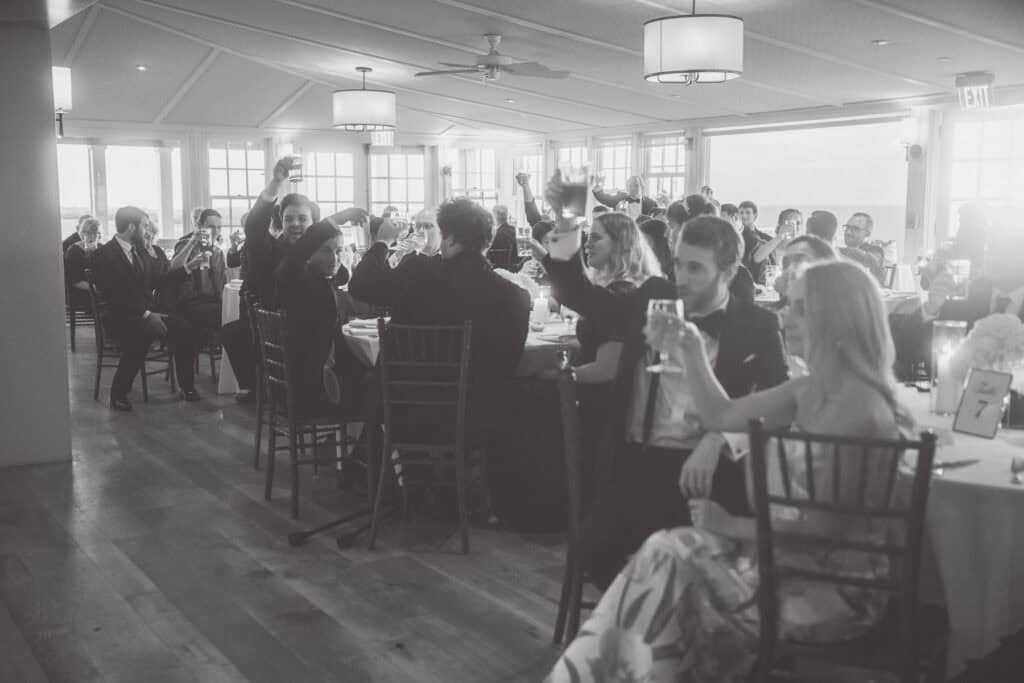 Wedding guests seated at tables in a bright banquet hall raise their glasses in a toast, captured beautifully by Nantucket Wedding Photographer Kaity Farrell.