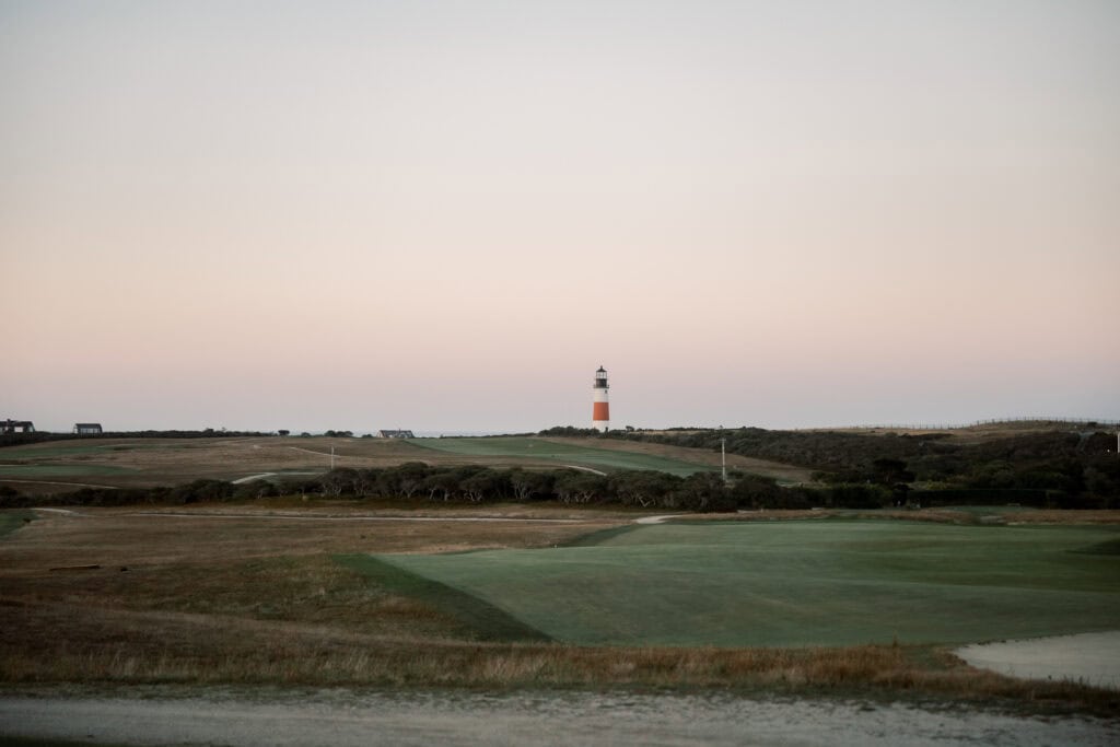A lighthouse stands on a distant hill overlooking Sankaty Head Golf Club’s grassy landscape at sunset, with a clear sky in the background—an idyllic scene for any wedding venue, captured beautifully by Nantucket Wedding Photographer Kaity Farrell.
