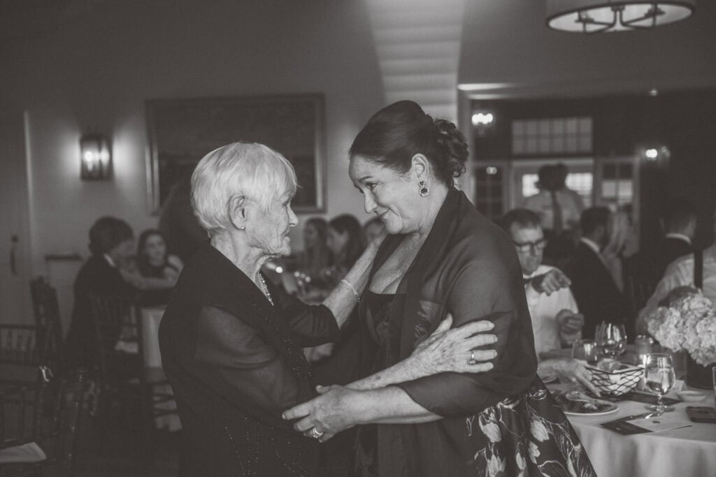 Two women stand facing each other, holding hands and smiling, in a formal dining setting with guests seated at tables—captured beautifully by Nantucket Wedding Photographer Kaity Farrell.