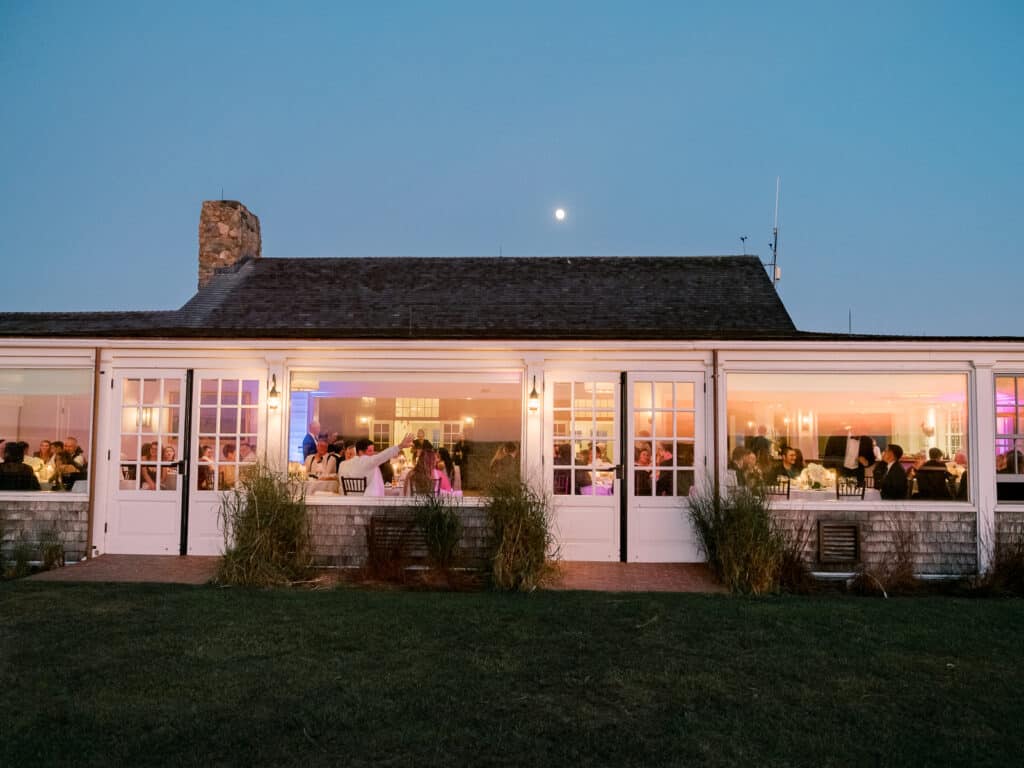 A view of a brightly lit event or reception inside a building with large windows at dusk, captured by Nantucket Wedding Photographer Kaity Farrell, with people seated at tables and a full moon glowing in the sky.