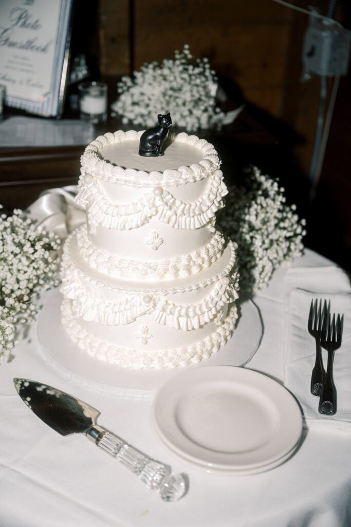 A three-tiered white wedding cake decorated with ruffles and topped with a black cat figurine sits on a table, beautifully captured by Nantucket Wedding Photographer Kaity Farrell, surrounded by plates, cutlery, and a cake server.