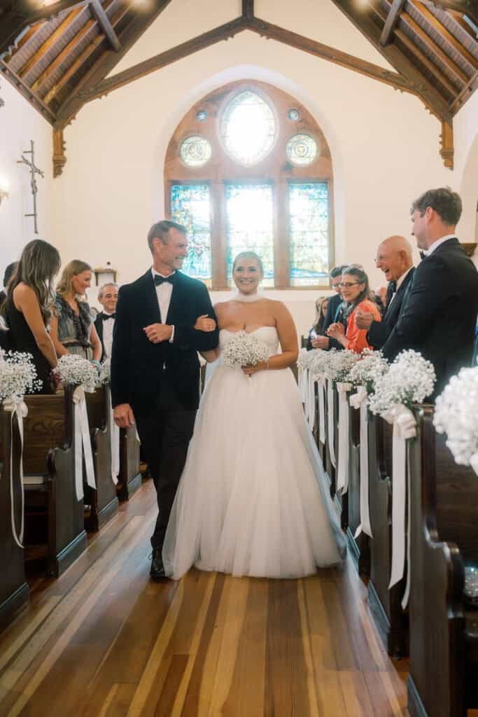 A bride in a white gown walks down the aisle with an older man in a suit, surrounded by seated guests in a church decorated with white flowers and ribbons.