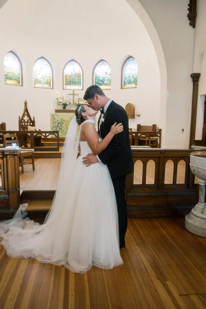 A bride and groom kiss inside a church, both dressed in formal wedding attire, standing on a wooden floor with stained glass windows in the background.
