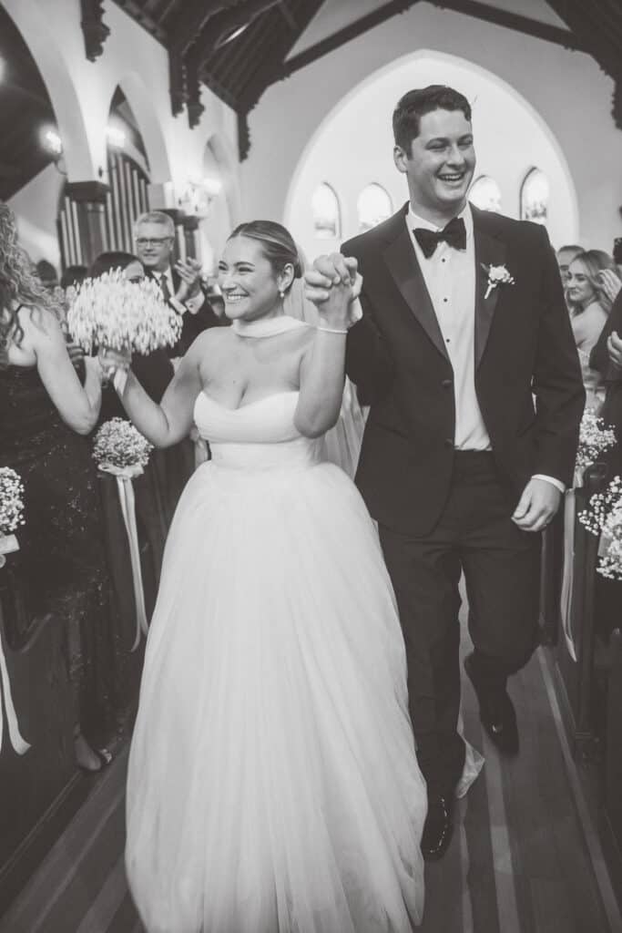 A bride and groom walk down the aisle in a church, smiling and holding hands, surrounded by guests—captured beautifully by Nantucket Wedding Photographer Kaity Farrell.