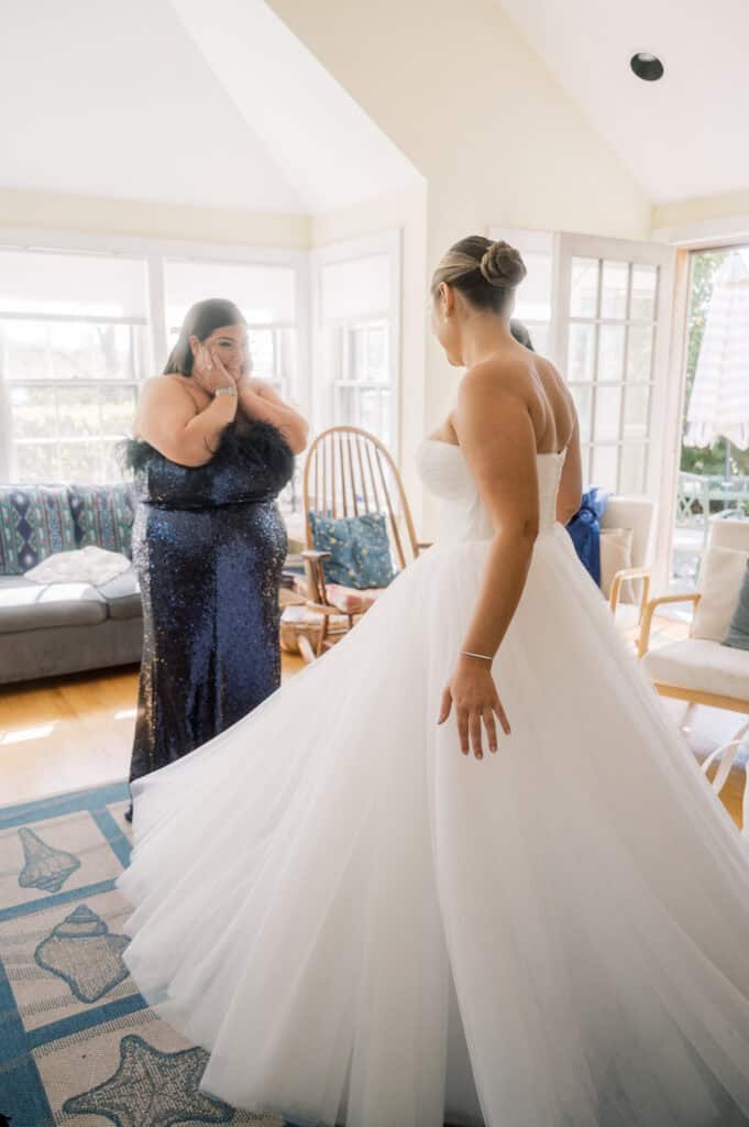 A woman in a white wedding dress stands in a bright room while another woman in a dark blue dress reacts emotionally, covering her mouth with her hands.