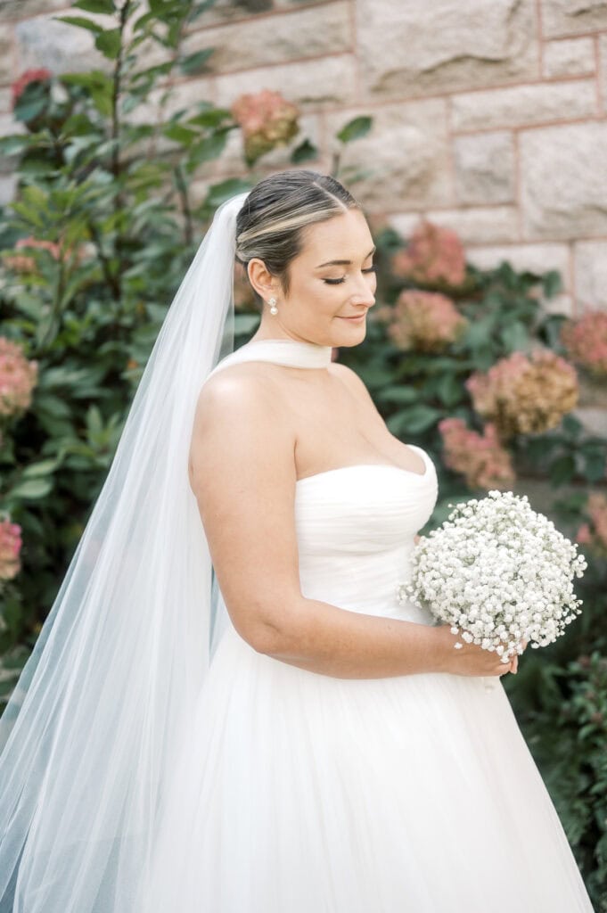 A bride in a strapless white gown and long veil holds a bouquet of baby's breath, standing near pink hydrangeas and a stone wall, beautifully captured by Nantucket wedding photographer Kaity Farrell.