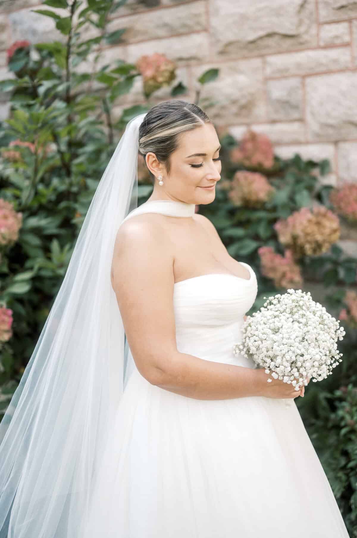 A bride in a strapless white wedding dress holds a bouquet of baby's breath flowers, standing outdoors near a stone wall and greenery, captured beautifully by Nantucket Wedding Photographer Kaity Farrell.