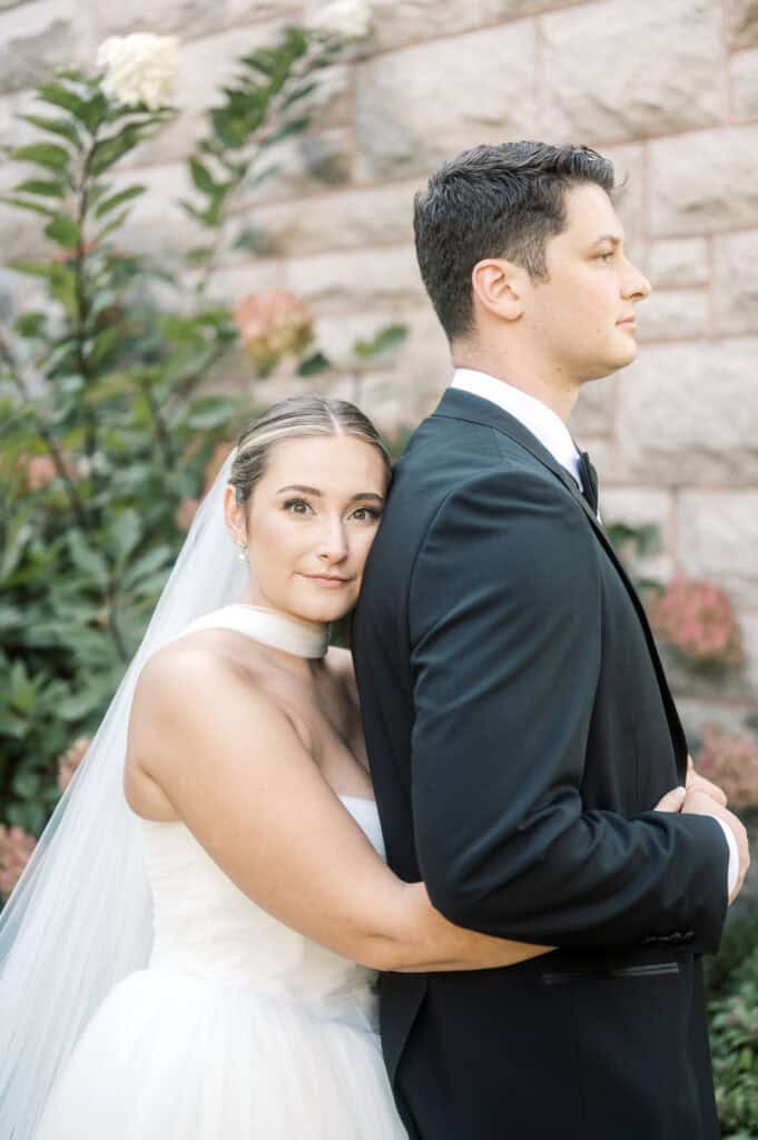 A bride and groom share a warm embrace outdoors against a stone wall, surrounded by lush greenery—capturing the timeless romance of a Nantucket wedding.