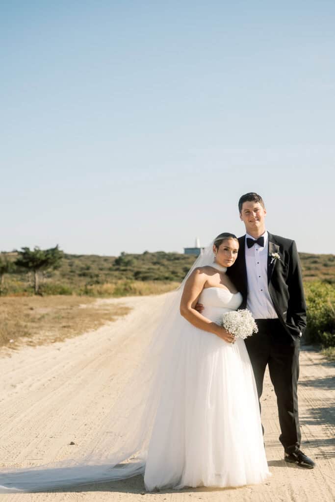 A bride in a white strapless gown and veil stands beside a groom in a black tuxedo on a sandy path with greenery, beautifully captured by Nantucket wedding photographer Kaity Farrell under a clear sky.