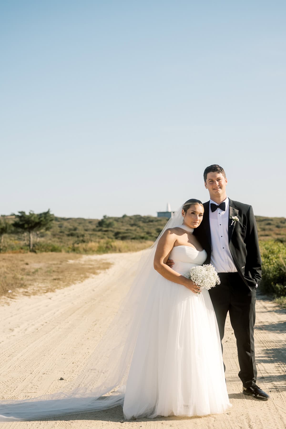 A bride in a white dress and a groom in a black suit stand together on a sandy path outdoors, with greenery and clear sky in the background, beautifully captured by Nantucket Wedding Photographer Kaity Farrell.