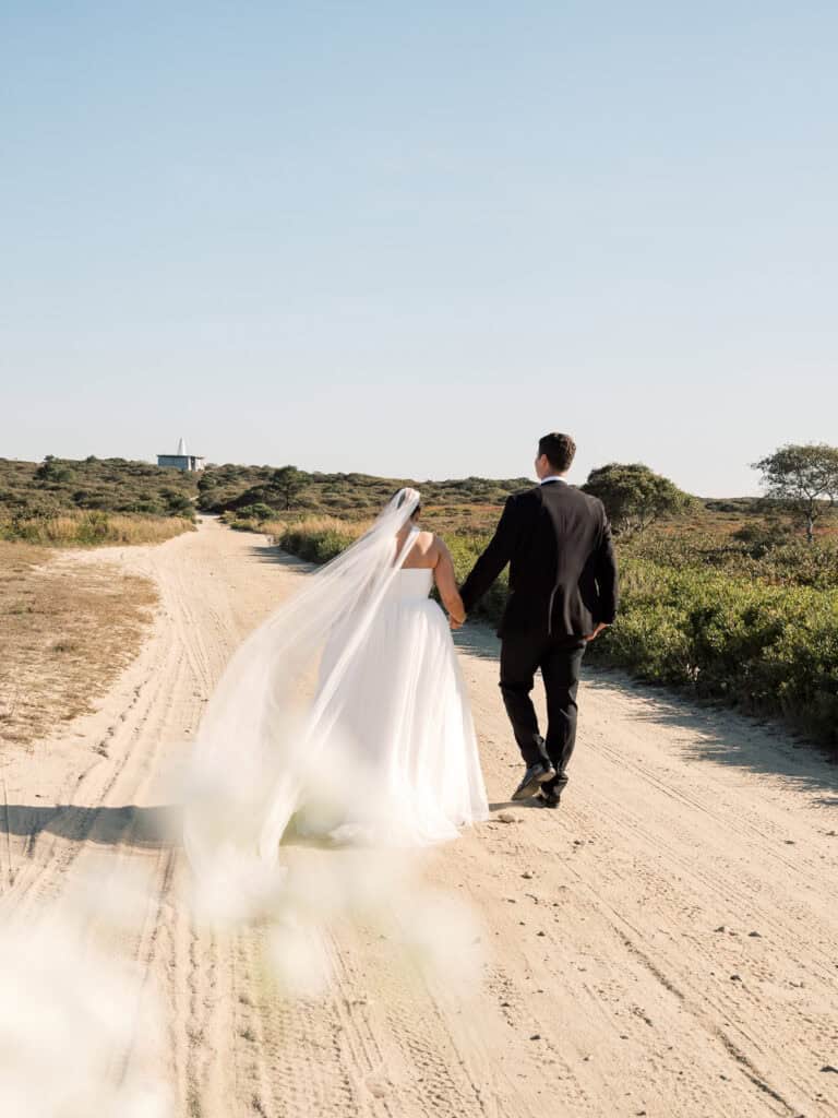A bride and groom walk hand in hand down a sandy path through a natural landscape, with the bride’s veil flowing behind her—captured beautifully by Nantucket wedding photographer Kaity Farrell.