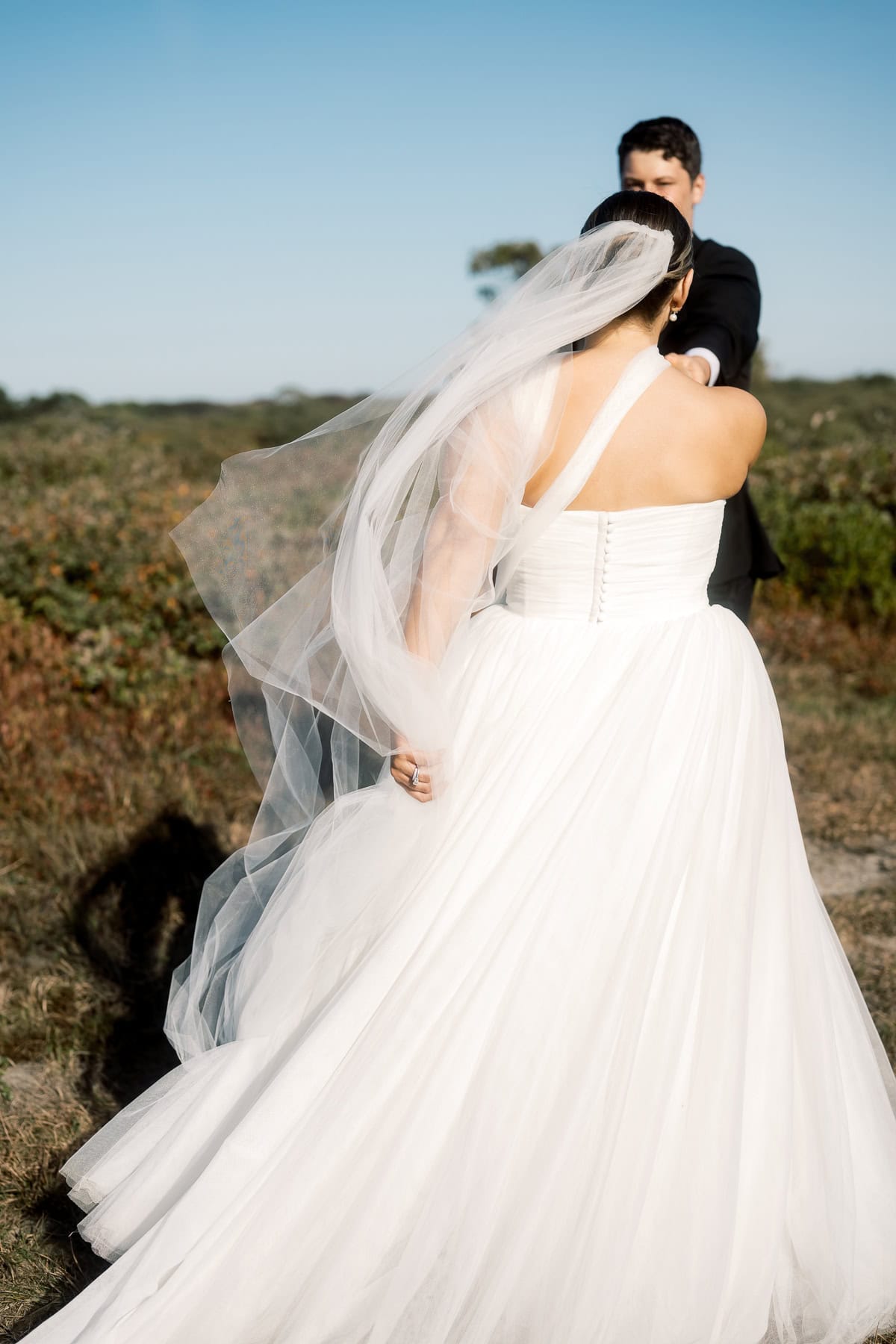 A bride in a white gown and veil walks outdoors, holding hands with a groom in a black suit on a sunny day, beautifully captured by Nantucket Wedding Photographer Kaity Farrell.