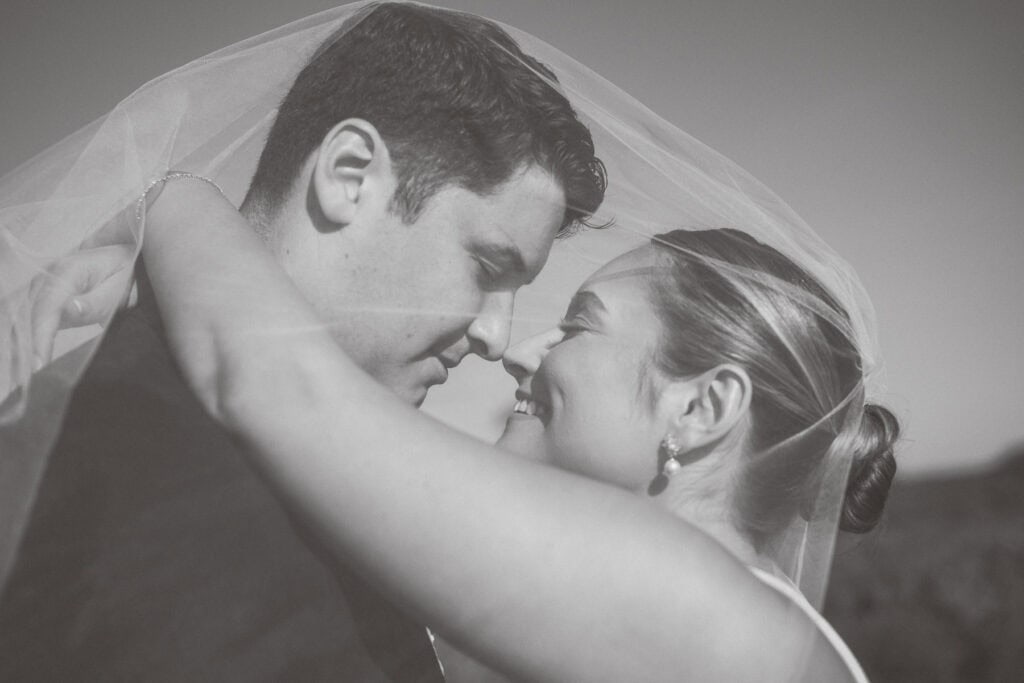 A bride and groom embrace closely, touching foreheads and smiling under a wedding veil in an outdoor setting, beautifully captured by Nantucket Wedding Photographer Kaity Farrell.