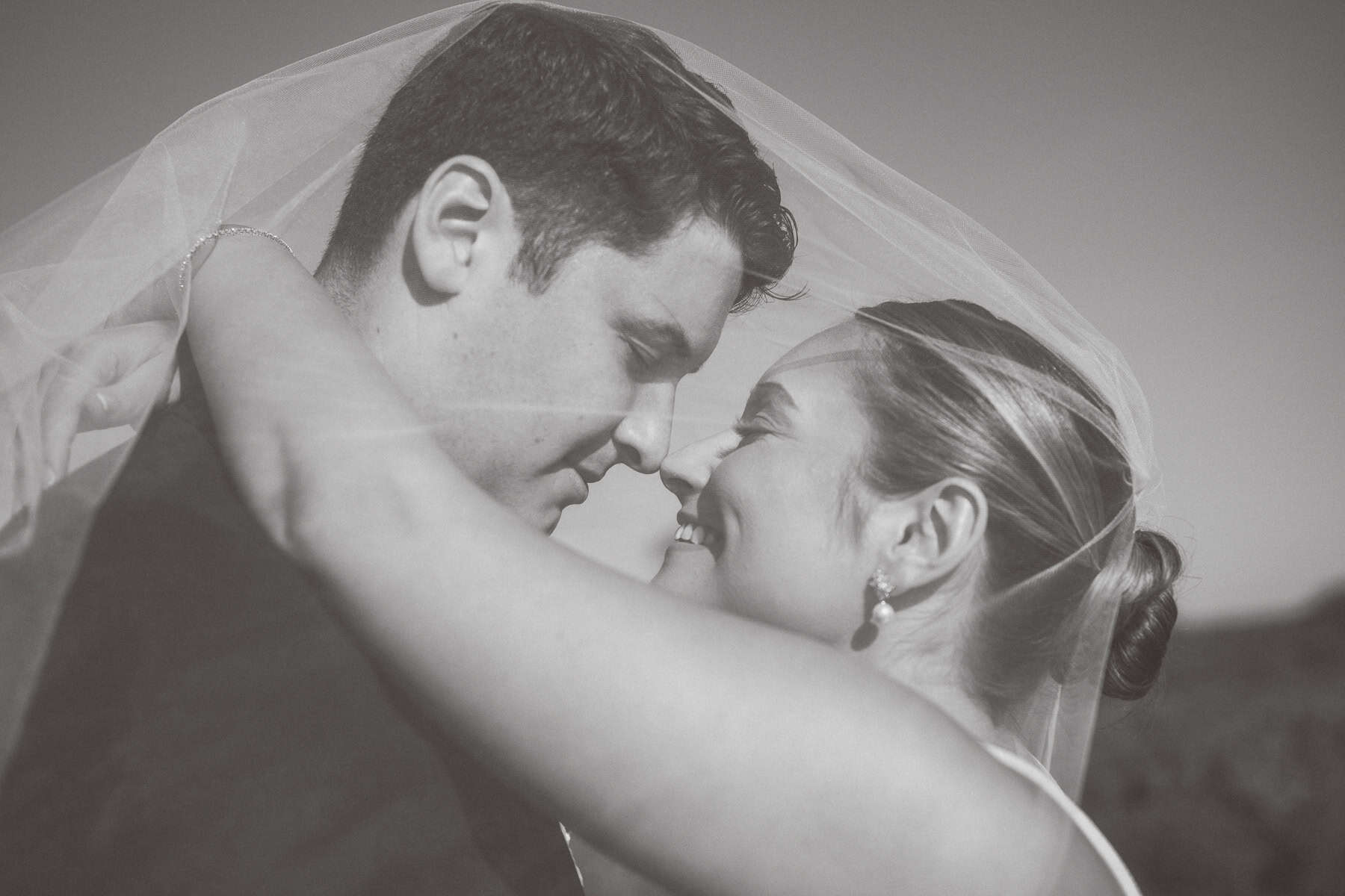 A bride and groom embrace closely, touching foreheads and smiling under a wedding veil in an outdoor setting, beautifully captured by Nantucket Wedding Photographer Kaity Farrell.