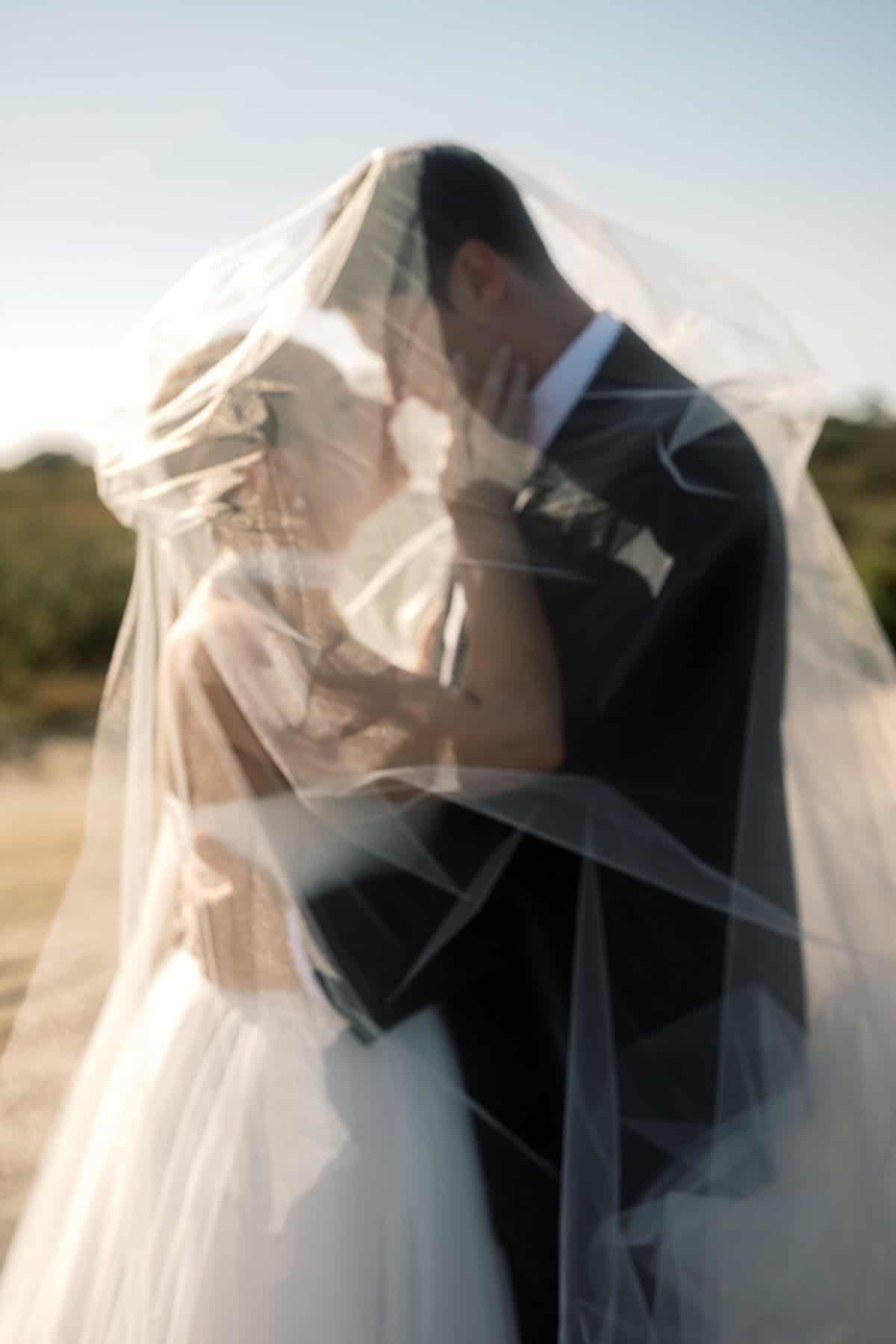 A bride and groom stand close together outdoors, embracing under a sheer white veil with soft natural light, captured beautifully by Nantucket Wedding Photographer Kaity Farrell.