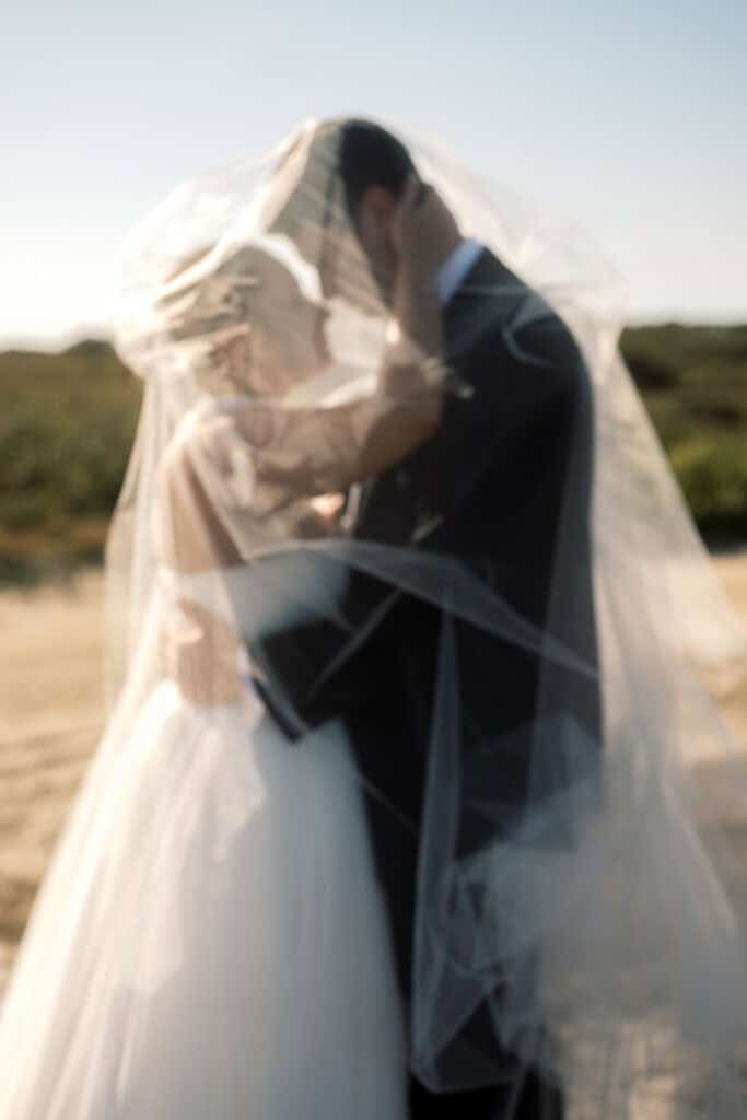 A bride and groom stand outdoors, embracing under a sheer wedding veil with natural scenery in the background, beautifully captured by Nantucket wedding photographer Kaity Farrell.