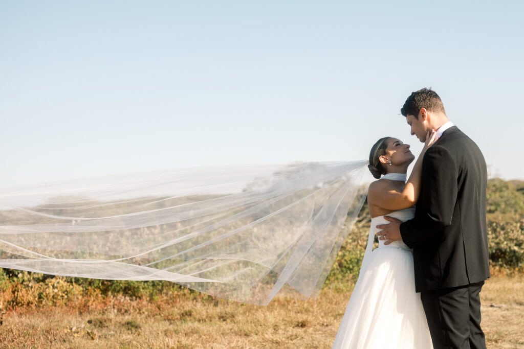 A bride and groom embrace outdoors during their Nantucket wedding, the bride’s veil flowing in the wind against a clear sky and grassy landscape.