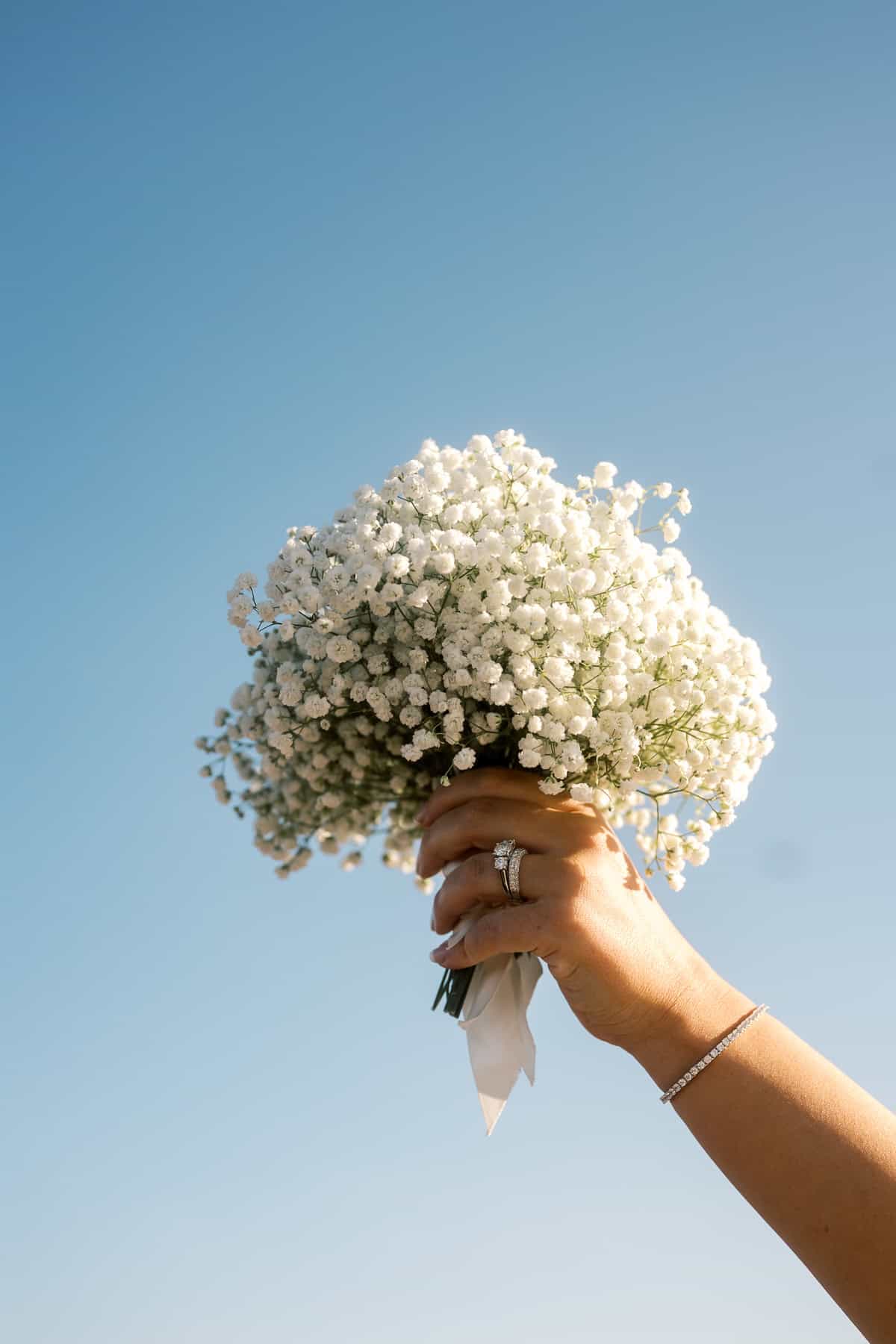 A hand holding a bouquet of small white flowers against a clear blue sky, adorned with a ring and bracelet—captured beautifully by Nantucket Wedding Photographer Kaity Farrell.