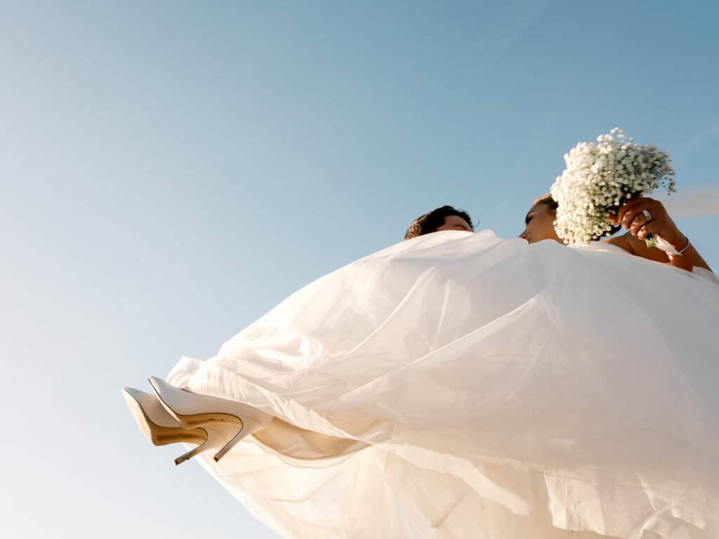 Low-angle view of a bride in a white dress holding a bouquet, with the sky in the background and her white heels visible—captured by Nantucket Wedding Photographer Kaity Farrell.