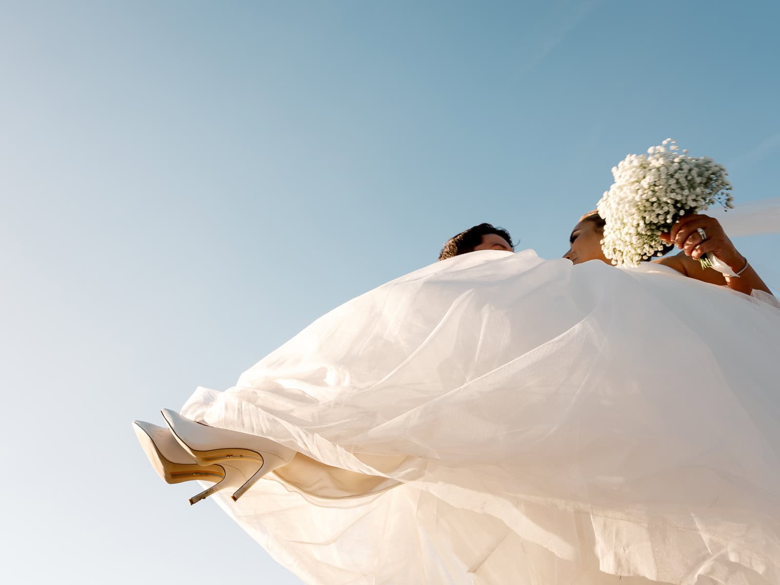 Low-angle view of a bride in a white dress holding a bouquet, with the sky in the background and her white heels visible—captured by Nantucket Wedding Photographer Kaity Farrell.