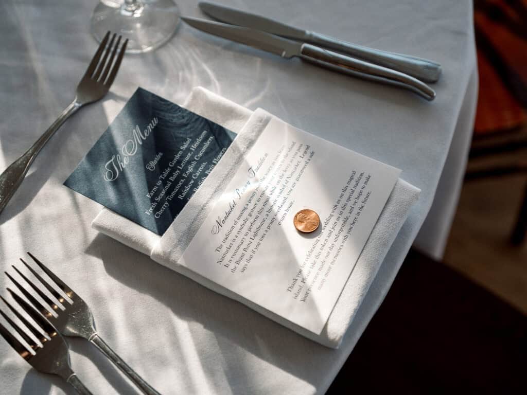 A white napkin on a table holds a menu, an event card, and a penny, with silverware and a wine glass nearby—captured beautifully by Nantucket Wedding Photographer Kaity Farrell.