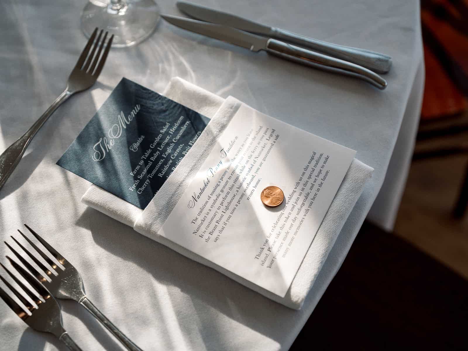 A white napkin on a table holds a menu, an event card, and a penny, with silverware and a wine glass nearby—captured beautifully by Nantucket Wedding Photographer Kaity Farrell.