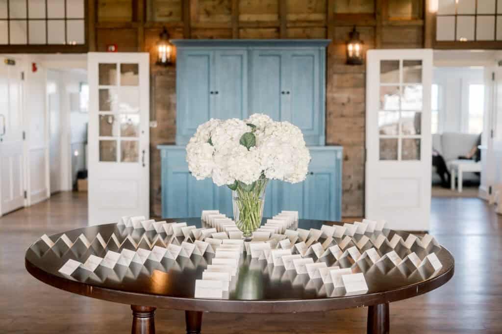 A round table with a vase of white hydrangeas at the center, surrounded by rows of place cards, in a bright room with double doors and a blue cabinet, photographed by Nantucket Wedding Photographer Kaity Farrell.