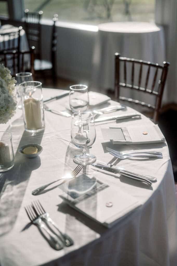 A round table set for a formal event with white tablecloth, glassware, cutlery, napkins, menus, candles, and wooden chairs in a sunlit room, beautifully captured by Nantucket Wedding Photographer Kaity Farrell.