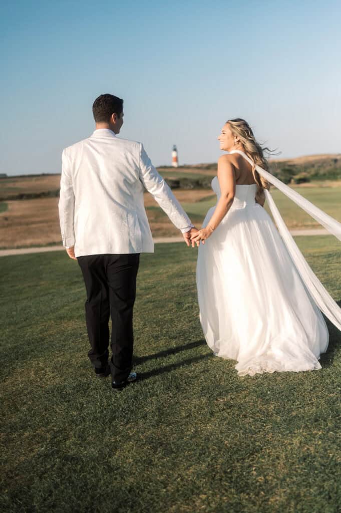 A bride in a white gown and a groom in a white jacket hold hands and walk on grass with a lighthouse in the background, captured beautifully by Nantucket Wedding Photographer Kaity Farrell.