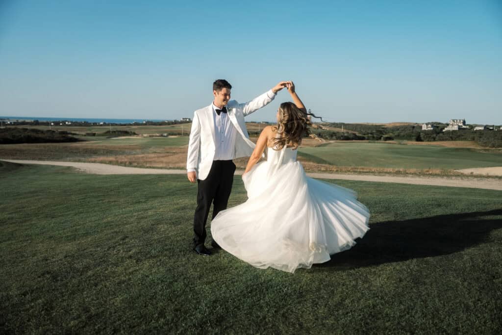A groom in a white jacket twirls a bride in a white gown on a grassy field under a clear sky, beautifully captured by Nantucket Wedding Photographer Kaity Farrell with the distant landscape as their backdrop.