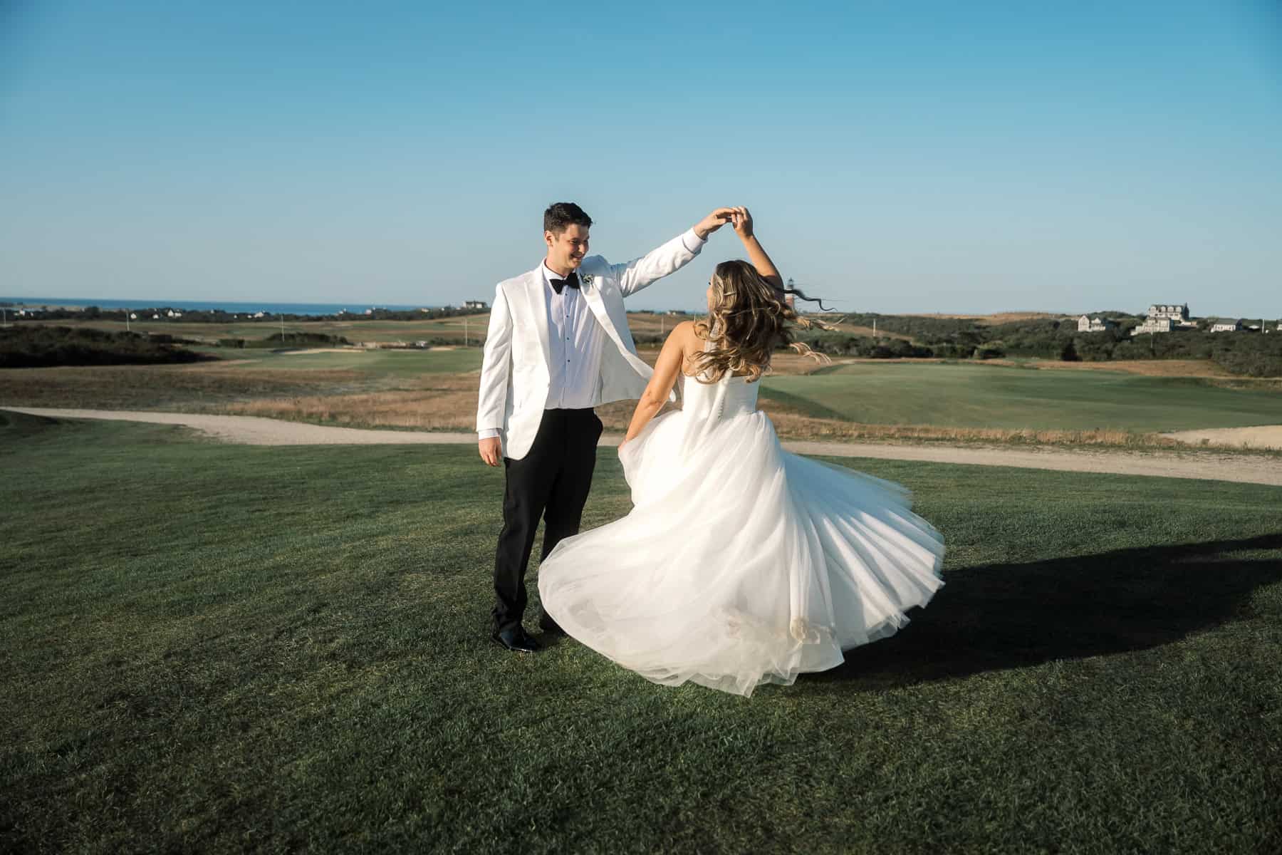 A groom in a white jacket twirls a bride in a white gown on a grassy field under a clear sky, beautifully captured by Nantucket Wedding Photographer Kaity Farrell with the distant landscape as their backdrop.