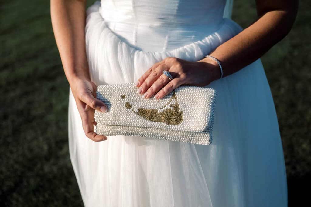 A person in a white dress holds a beaded clutch with a gold design, showing a ring and bracelet on their left hand—captured by Nantucket Wedding Photographer Kaity Farrell.