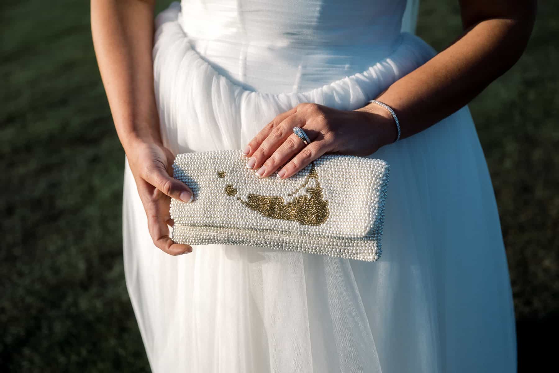 A person in a white dress holds a beaded clutch with a gold design, showing a ring and bracelet on their left hand—captured by Nantucket Wedding Photographer Kaity Farrell.