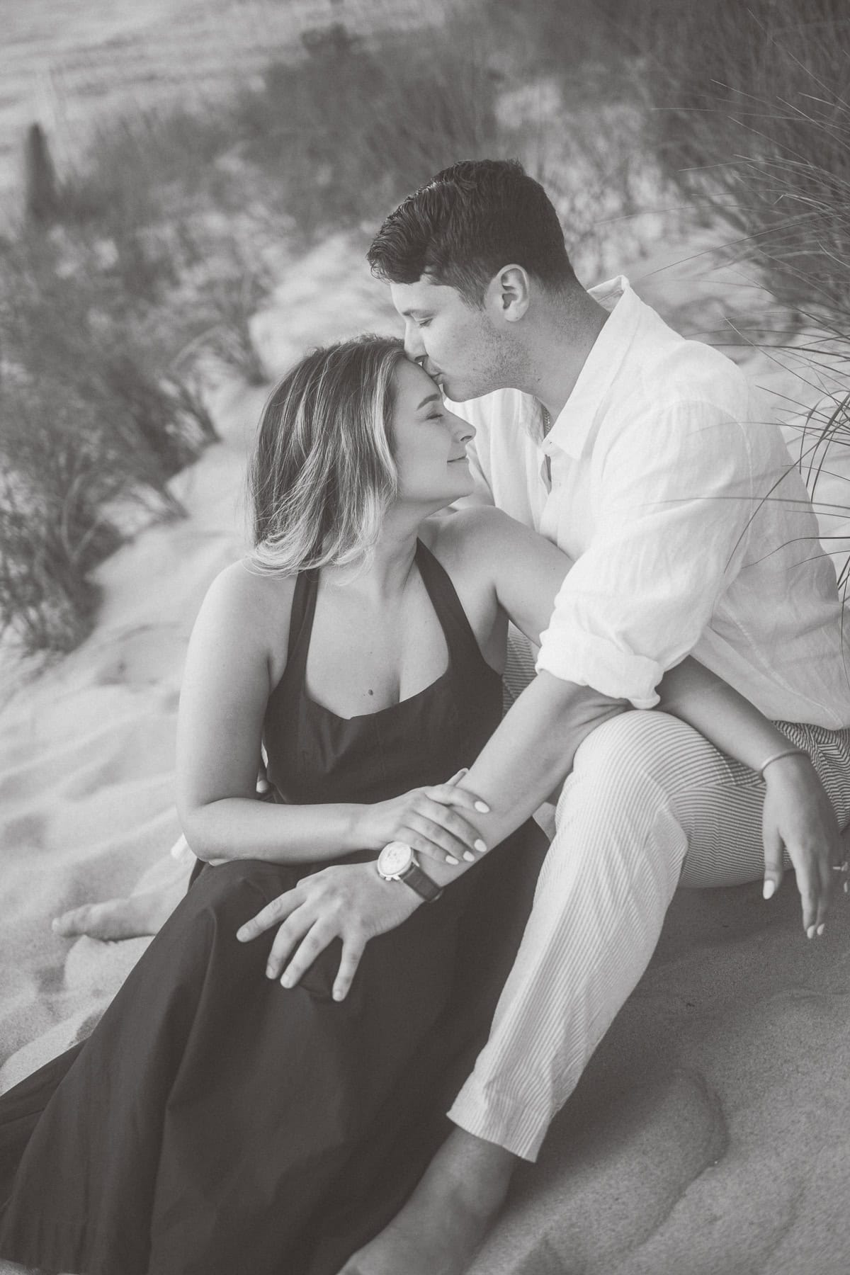 A Nantucket couple sits close together on a sandy beach; the man kisses the woman's forehead as she looks at him and holds his arm during their engagement photo session with Nantucket photographer Kaity Farrell.