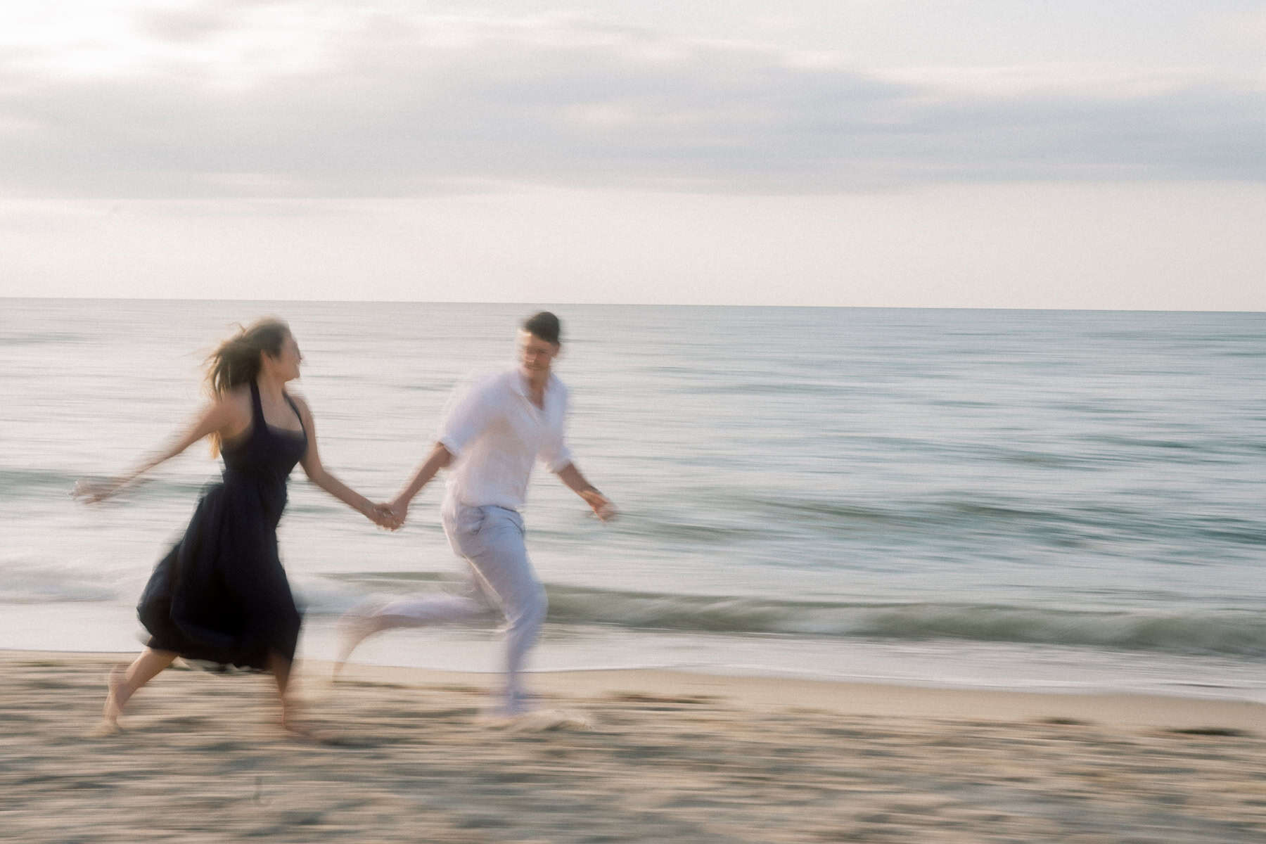 A Nantucket couple hold hands and run along a sandy beach near the ocean under a cloudy sky; Nantucket photographer Kaity Farrell captures this slightly blurred engagement photo session full of motion.