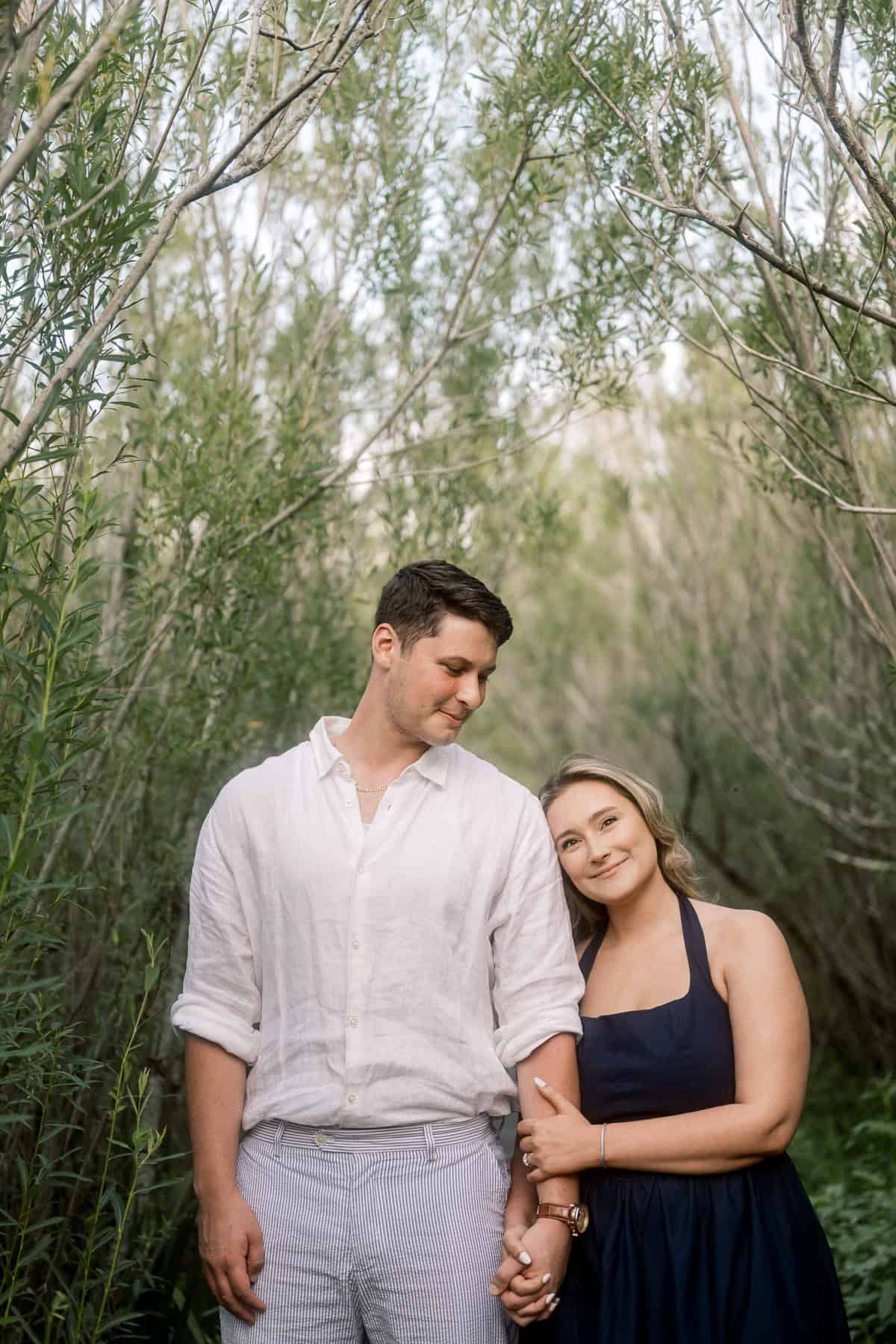 A man in a white shirt and a woman in a navy dress stand together in a wooded area, holding hands; the woman rests her head on his shoulder during their engagement photo session with Nantucket photographer Kaity Farrell.