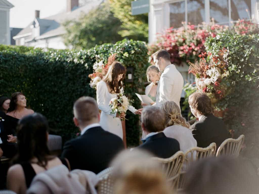 A couple stands together during an outdoor wedding ceremony, captured on film photo by Nantucket Wedding Photographer Kaity Farrell, with guests seated and lush greenery and floral arrangements in the background.