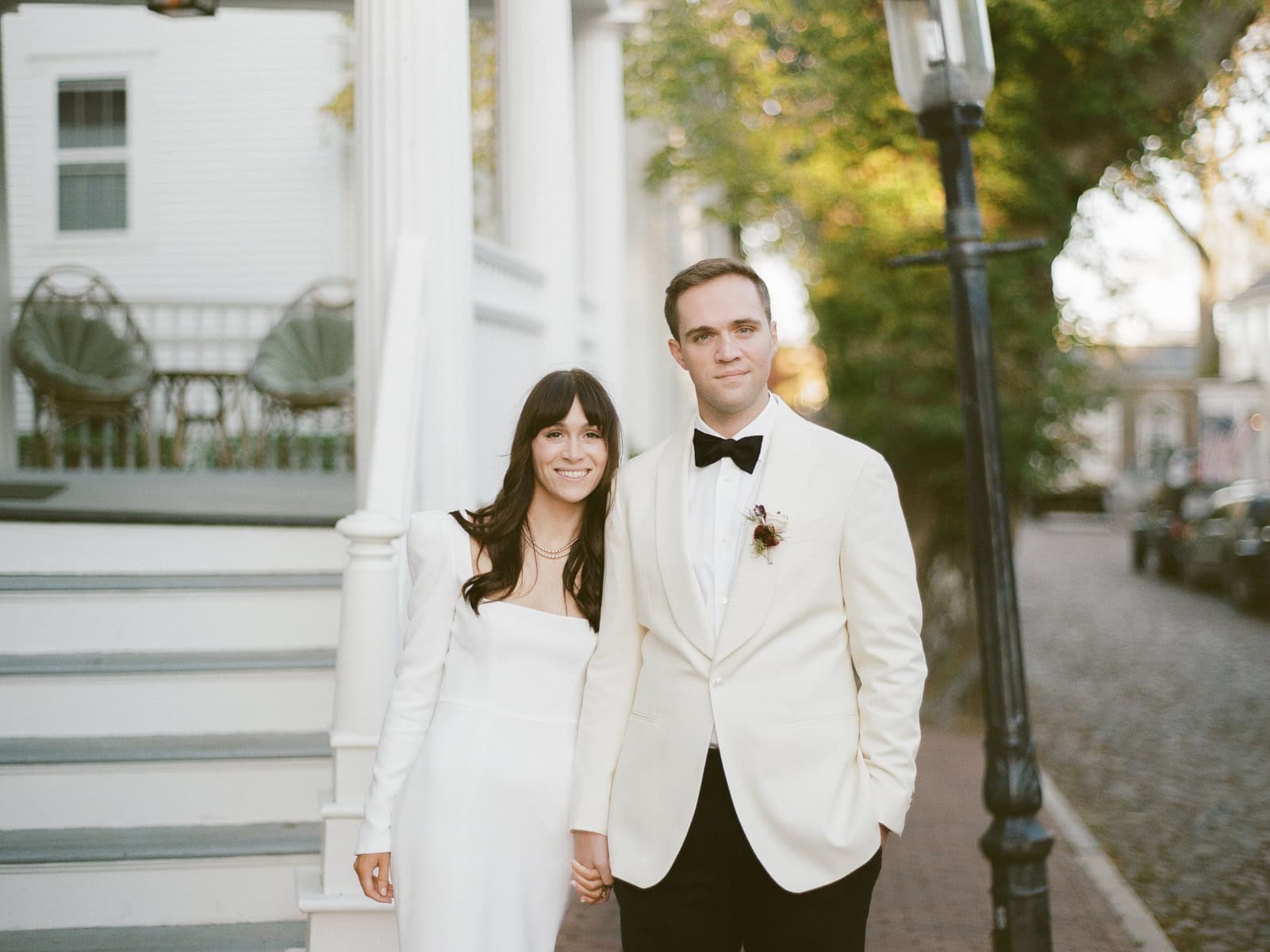 A couple in formal white attire stands hand in hand on a sidewalk by a staircase, captured in a timeless film photo by Nantucket Wedding Photographer Kaity Farrell. The man wears a tuxedo and the woman is in a long-sleeve dress.