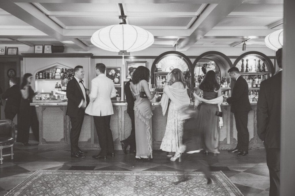 A group of people dressed in formal attire stand at a bar, engaging in conversation. Nantucket Wedding Photographer Kaity Farrell captures the indoors black and white scene, with a blurred figure in motion in the foreground.