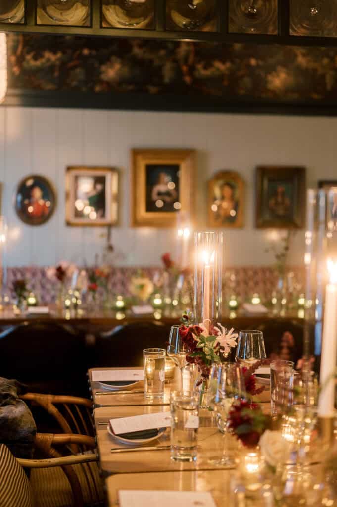 A warmly lit dining table is set with glassware, menus, flowers, and candles at Sister Ship restaurant; framed artwork hangs on the wall in the background at a Nantucket wedding photographed by Kaity Farrell.